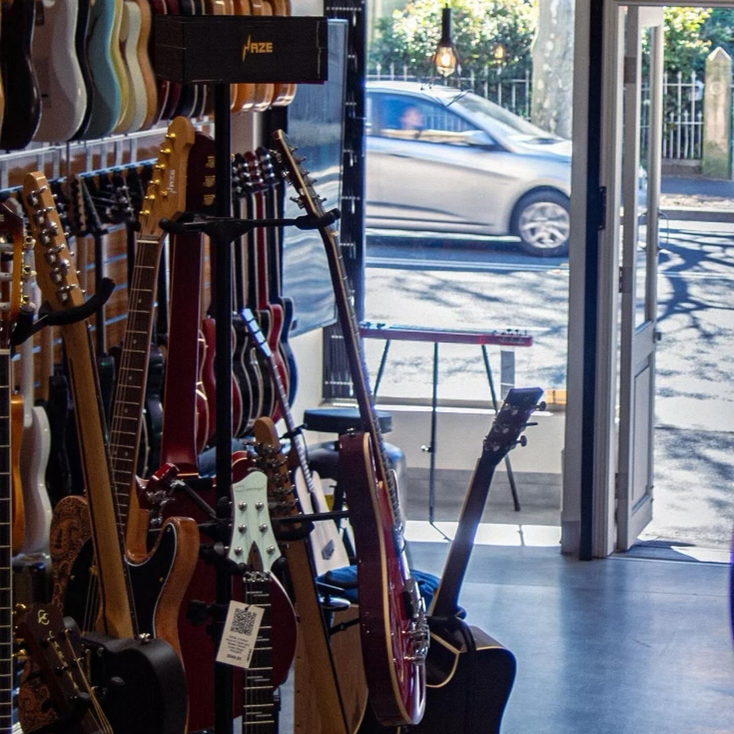 Guitar store interior with guitars on display and a car outside through the open door.